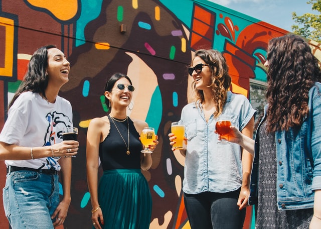 Four women laugh while holding drinks outside.