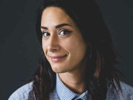 A brown-haired woman wearing a tie looks into the camera.