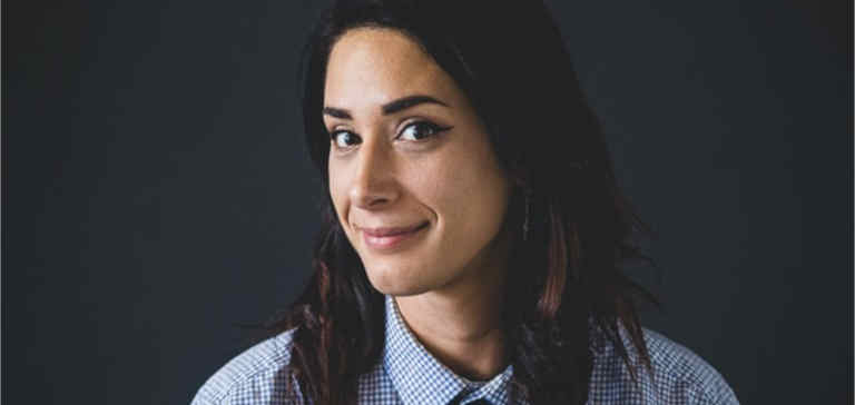 A brown-haired woman wearing a tie looks into the camera.