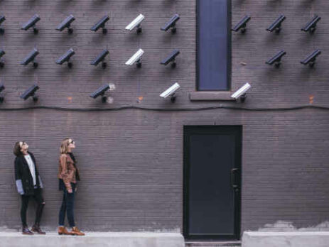 Two women look up at many black and white security cameras.