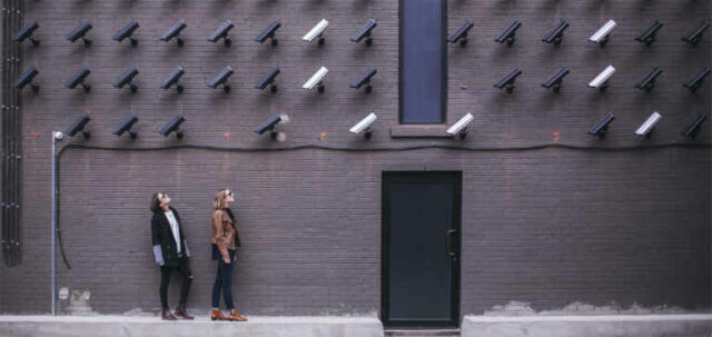 Two women look up at many black and white security cameras.