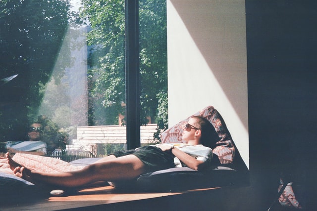 A man in a white t-shirt lies in the sunshine in a window seat.