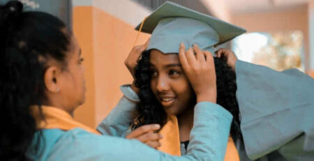 A woman helps a young graduate girl put on her cap and gown.