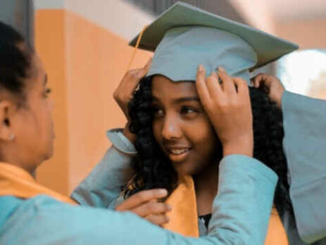 A woman helps a young graduate girl put on her cap and gown.