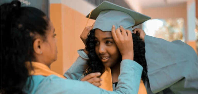 A woman helps a young graduate girl put on her cap and gown.