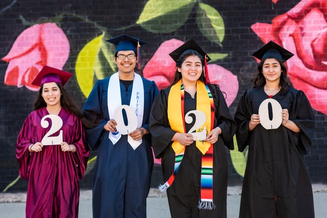 Four young graduates stand in front of a brick wall in their cap and gowns.