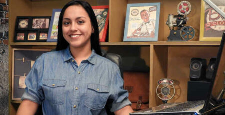 A woman in a blue shirt sits near a computer and some framed photos.