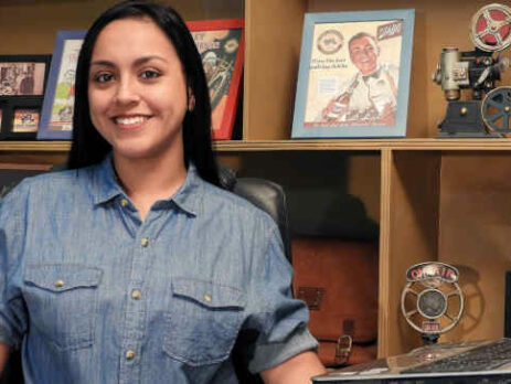 A woman in a blue shirt sits near a computer and some framed photos.