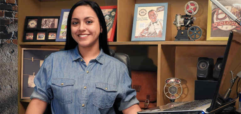 A woman in a blue shirt sits near a computer and some framed photos.