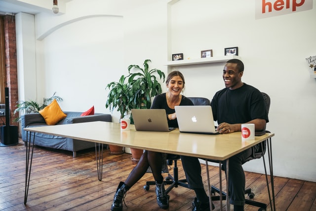 A man and woman laugh as they work together on two laptop computers.