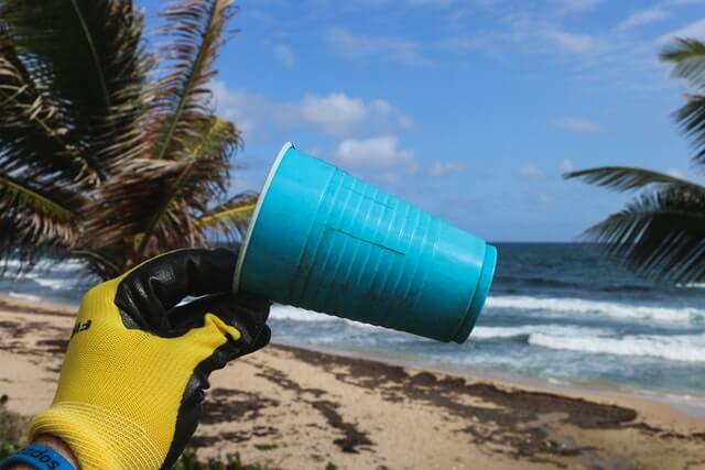 A gloved hand holds up a plastic yellow cup on the beach.