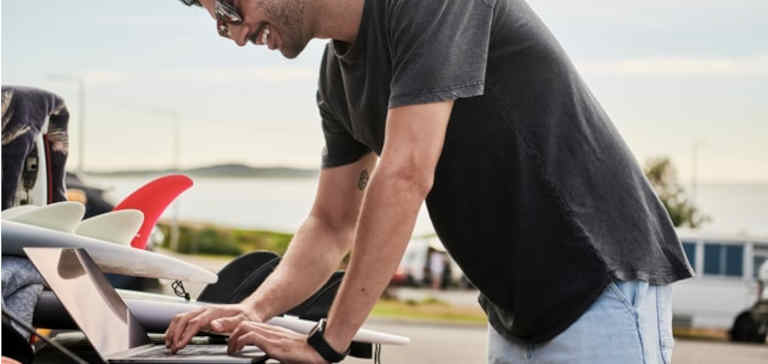 A man in a grey shirt works on a laptop computer outside.