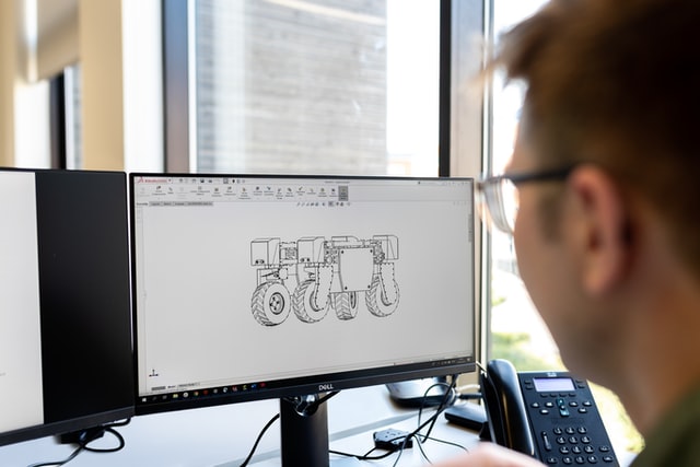 A man sits near a computer displaying a 3D rendering.