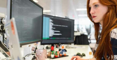 A redhead woman works on a computer with multiple monitors.