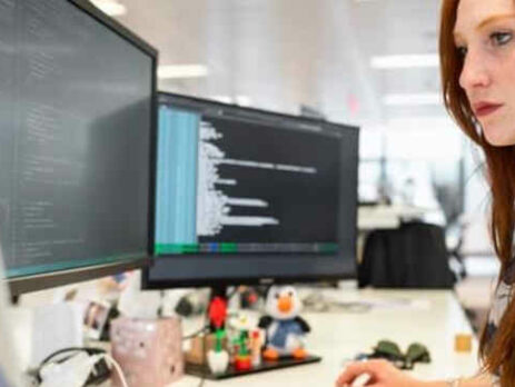 A redhead woman works on a computer with multiple monitors.