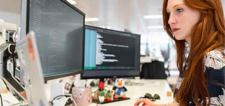A redhead woman works on a computer with multiple monitors.