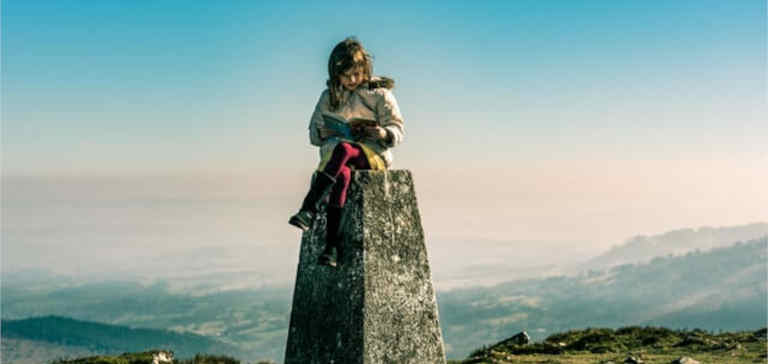 A girl reads a book on top of a pedestal on a mountain.