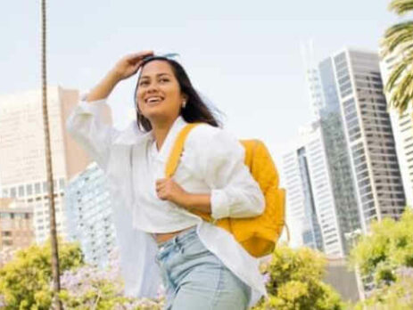 A woman wearing sunglasses and a yellow backpack walks outside.