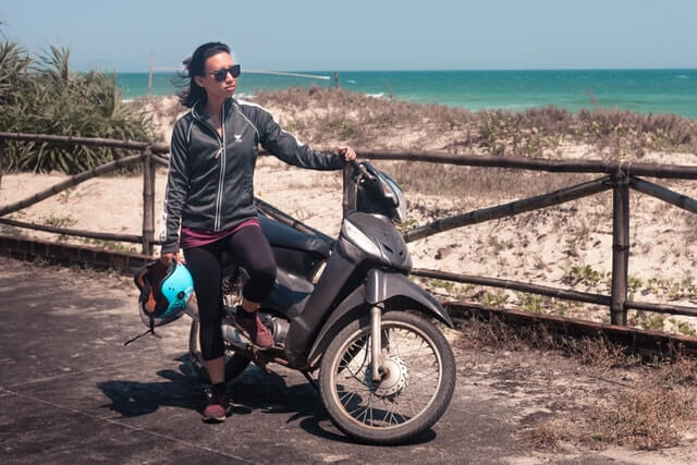 A woman stands near a motorbike holding a helmet.