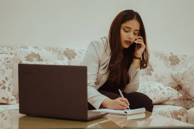 A woman in a white blazer writes and talks on the phone near a laptop.