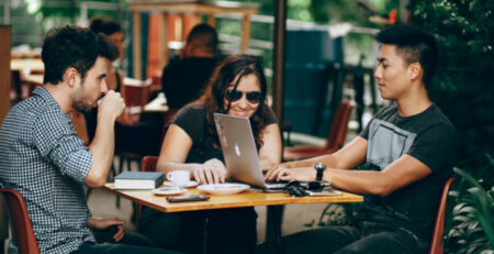 Two men and a woman sit outside at a cafe and work on a laptop.