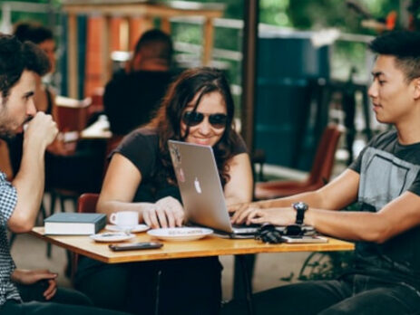 Two men and a woman sit outside at a cafe and work on a laptop.