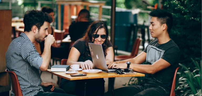 Two men and a woman sit outside at a cafe and work on a laptop.