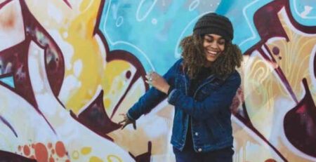 A woman wearing a hat waves her arms in front of some graffiti.