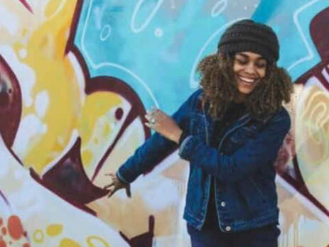 A woman wearing a hat waves her arms in front of some graffiti.