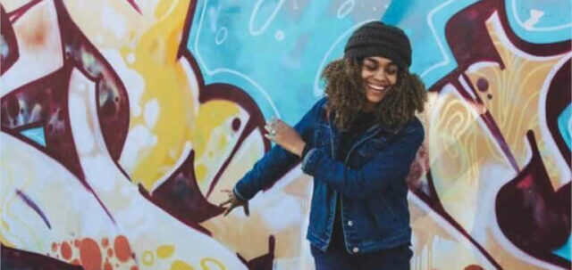 A woman wearing a hat waves her arms in front of some graffiti.