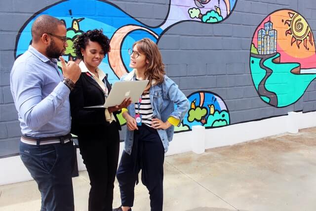 Three people talk around a computer in front of graffiti.