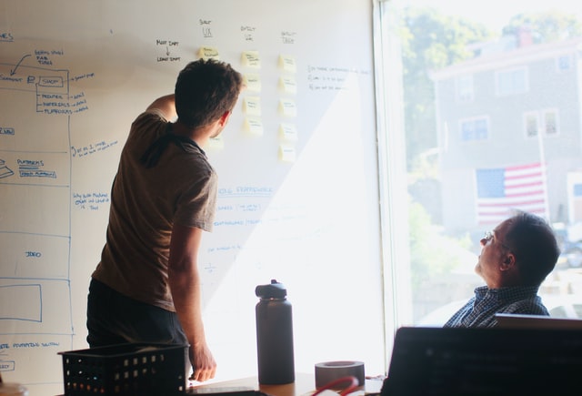 Two men talk while gesturing toward information on a whiteboard.