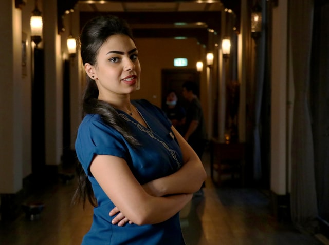 A woman wearing a blue t-shirt stands with her arms folded across her chest.