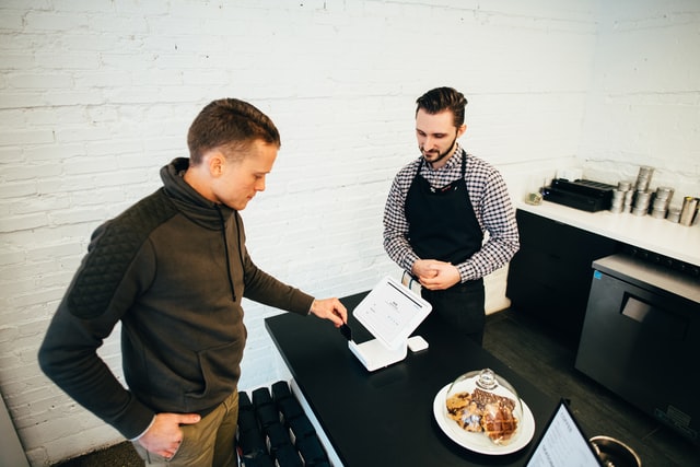 Two men speak over a small white cash register.