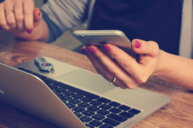 A person holding a smartphone near a laptop computer.