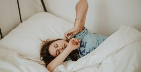 A woman in a blue t-shirt stretches as she wakes up in the morning.