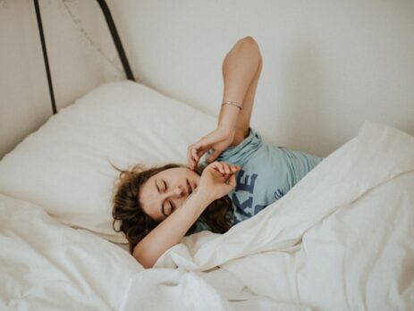 A woman in a blue t-shirt stretches as she wakes up in the morning.