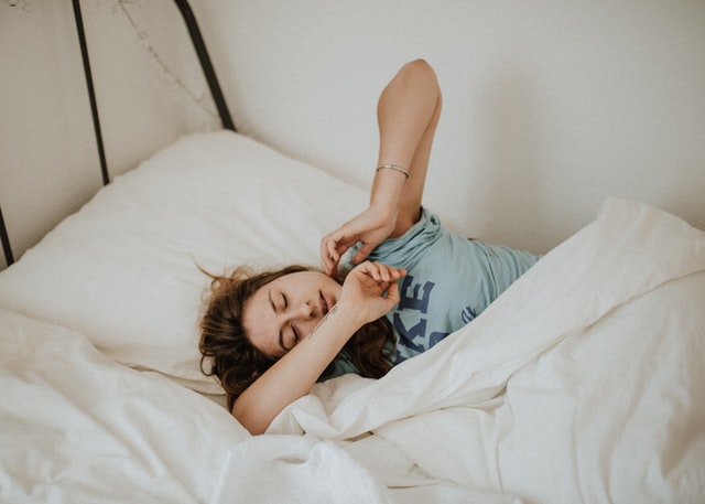 A woman in a blue t-shirt stretches as she wakes up in the morning.