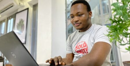 A man sits at a white table and works on a laptop computer.