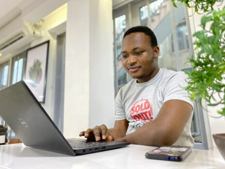 A man sits at a white table and works on a laptop computer.