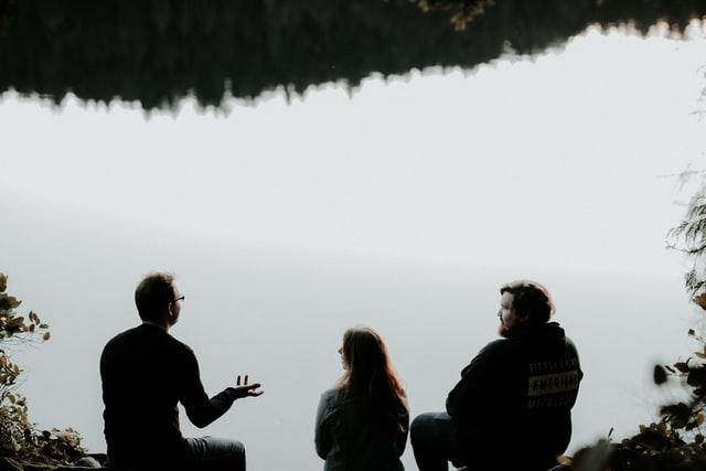 Three people sit near a lake and talk.