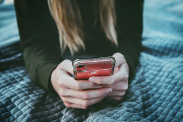 Hands operate a smartphone while a person lies across the bed.