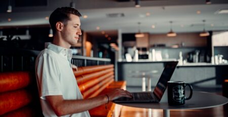 A man types on a laptop in a cafe with a window