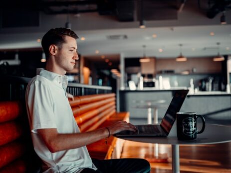 A man types on a laptop in a cafe with a window