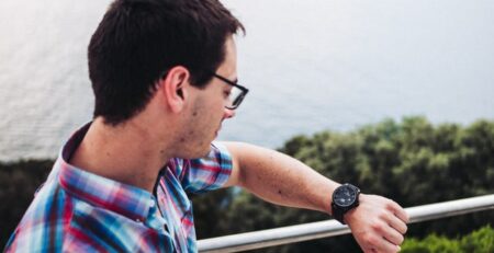 A man in a colorful checkered shirt looks at his watch near the ocean.