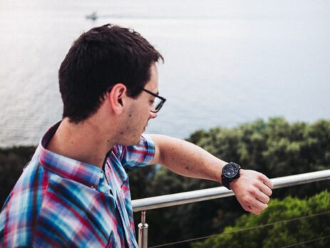 A man in a colorful checkered shirt looks at his watch near the ocean.