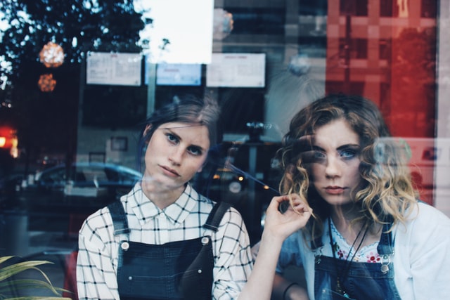 Two girls stare at the camera with fed-up stares behind a glass shop window.
