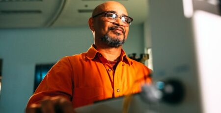 A man wearing glasses smiles while he works on a laptop.