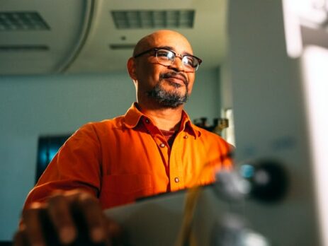 A man wearing glasses smiles while he works on a laptop.