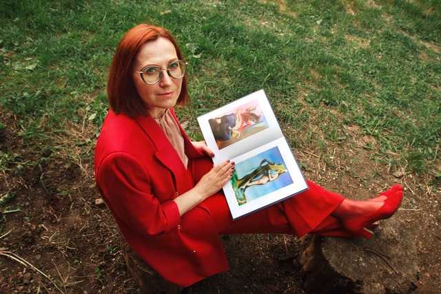 A woman in a red suit sits with a magazine on green grass.